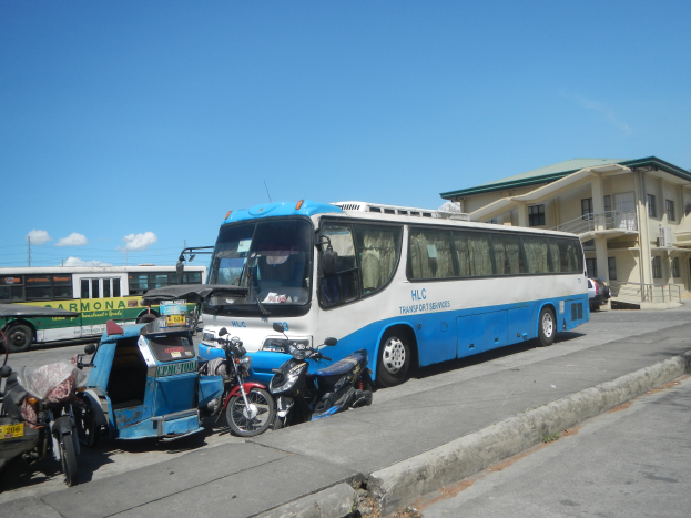Ein blauer und weißer Bus parkt am Straßenrand und ist von Motorrädern und anderen Fahrzeugen umgeben. Im Hintergrund sind Gebäude, Strommasten mit Drähten und ein bewölkter Himmel zu sehen.