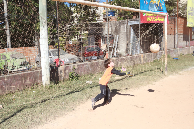 Ein junger Junge kickt einen Füüball in ein Tor auf einem Feld mit Gebäuden, Bäumen und Fahrzeugen im Hintergrund unter einem klaren blauen Himmel.