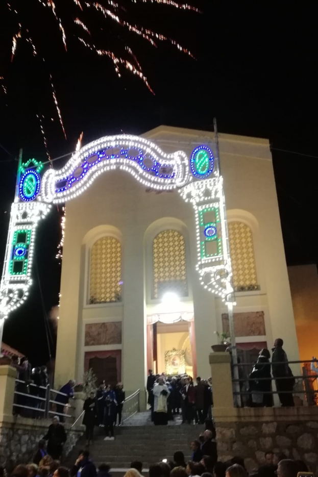 Eine Gruppe von Menschen, die vor einem Gebäude mit einem beleuchteten Bogengang, Treppen mit Geländern und Topfpflanzen steht und das Silvesterfeuerwerk im Hintergrund betrachtet.