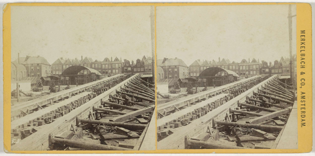Ein altes Schwarz-Weiß-Foto einer Bahnschiene mit Gebäuden im Hintergrund, das den Text "Merkelbach & Co. Amsterdam" auf der rechten Seite und verstreute hölzerne Objekte im Vordergrund zeigt.