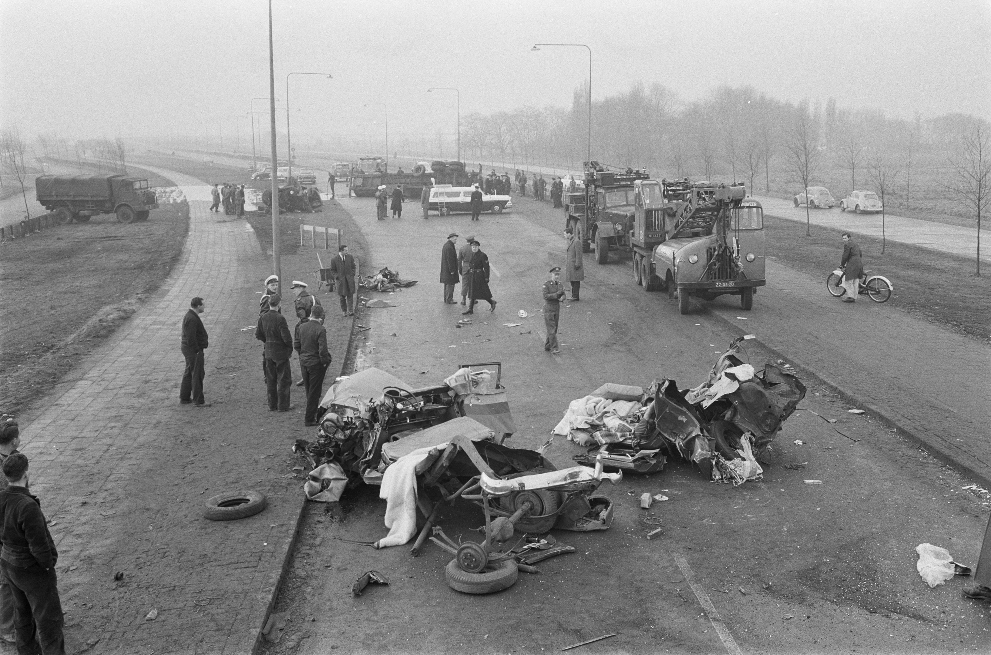 Schwarze und weiße Szene eines Autounfalls am Straßenrand mit mehreren Fahrzeugen, einer Gruppe von Menschen in der Nähe, Laternenpfählen, Bäumen und Himmel im Hintergrund.