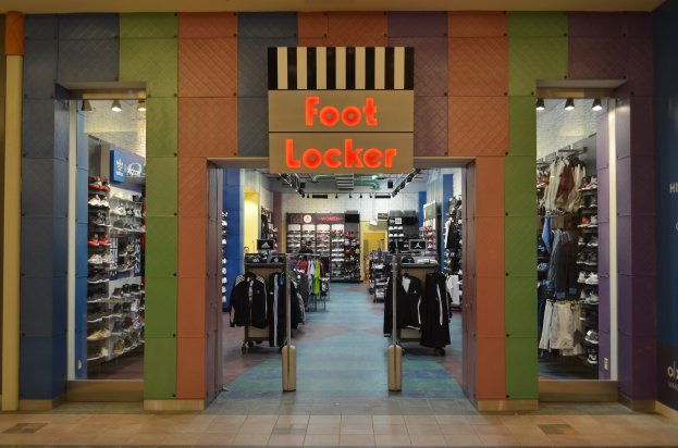 Innenansicht eines Foot Locker-Schuhgeschäfts mit Kleidung an Bügeln und Ständern unter Deckenbeleuchtung, mit sichtbarem Geschäftsfußboden.