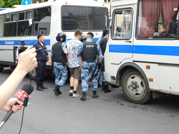Eine Gruppe von Polizeibeamten steht vor einem weißen und blauen Bus auf einer Straße mit Bäumen, Gebäuden und einem Schild, während eine Person in der Nähe ein Mikrofon und eine Kamera hält.