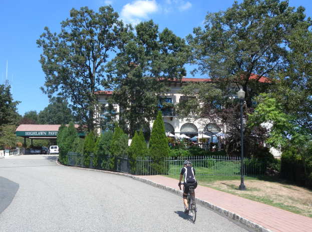 Eine Person mit Helm fährt ein Fahrrad auf einer von Bäumen gesäumten Straße mit Laternen, Gelöndern und Gras, mit einem Gebäude, einem Schuppen und einem bewölktem Himmel im Hintergrund.