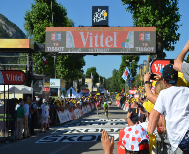 Radfahrer auf einer Straße unter einem Bogen mit der Aufschrift "Tour de France 2016", mit Zuschauern auf beiden Seiten und Bäumen, einem Hügel und einem klaren blauen Himmel im Hintergrund.