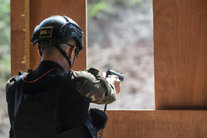 Ein Mann in Tarnkleidung, schwarzer Jacke und Helm zielt mit einer Pistole auf ein Ziel mit einem unscharfen Hintergrund.