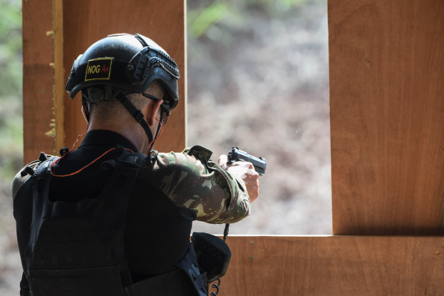 Ein Mann in Tarnkleidung, schwarzer Jacke und Helm zielt mit einer Pistole auf ein Ziel mit einem unscharfen Hintergrund.