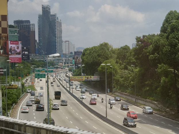 Eine belebte Autobahn mit hohen Gebäuden, Straßenlaternen, Lichtern, Werbetafeln, Bäumen und bewölktem Himmel.