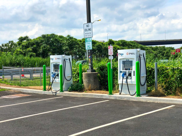 Elektrofahrzeug-Ladestation auf einem Parkplatz mit umgebender Infrastruktur, Vegetation und einer Brücke im Hintergrund.