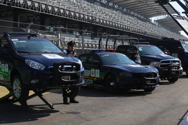 Polizeiautos vor einer Rennstrecke mit uniformierten Personen, ein Stadion mit Geländern und Maschendrahtzaun und Himmel im Hintergrund.