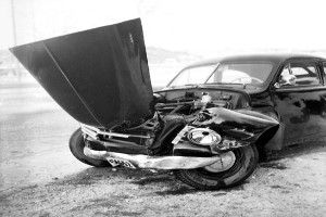 Ein Schwarz-Weiß-Foto eines schwer beschädigten Autos am Straßenrand mit eingedellter Motorhaube, zersplitterten Fenstern und öffnender Fahrertür, die den Motor freigibt.