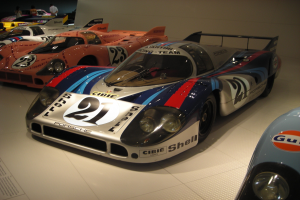 Ein Porsche 917K Le Mans Rennauto wird in einem Museum ausgestellt, umgeben von anderen Sportwagen und Menschen, mit einem textgefüllten Papier auf der linken Seite.