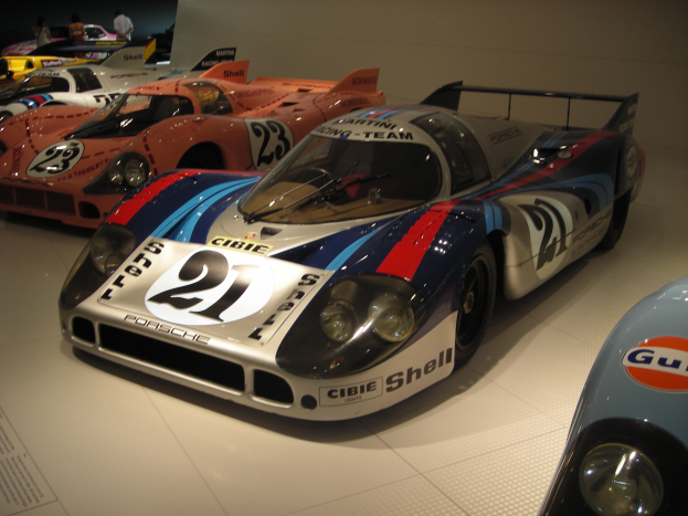 Ein Porsche 917K Le Mans Rennauto wird in einem Museum ausgestellt, umgeben von anderen Sportwagen und Menschen, mit einem textgefüllten Papier auf der linken Seite.