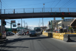 Eine belebte Stadtstraße mit Autos, Bussen, Motorrädern, einer Brücke mit Geländern und Laternen, Strommasten mit Drähten, Texttafeln und einem bewölkten Himmel.