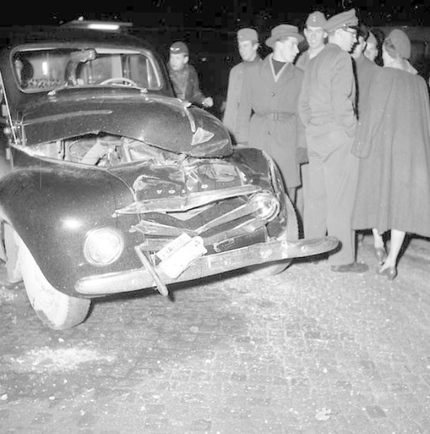 Schwarz-weißes Bild eines schwer beschädigten Autos am Straßenrand mit einer Gruppe von Menschen, die die Szene begutachten.