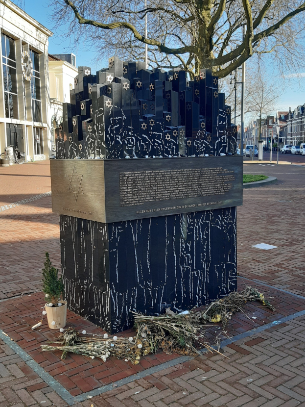 Denkmal in einem Stadtplatz für Holocaust-Opfer, mit einem Baum davor, einem Topf mit einer Pflanze, verstreuten trockenen Blättern und Gebäuden, Fahrzeugen und Bäumen im Hintergrund.