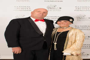 Ein Mann in einem schwarzen Anzug mit weißem Hemd und schwarzer Fliege steht neben einer Frau in einem eleganten Kleid und einem gemusterten Schal auf einem roten Teppich, mit einem weißen Banner mit Text im Hintergrund.