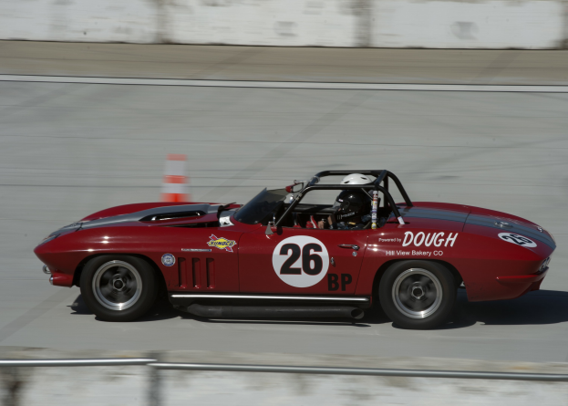 Roter Sportwagen mit einem Helmträger auf einer Rennstrecke, mit Text und Zahlen auf dem Auto, einem Verkehrskegel im Hintergrund und einem Eisenzaun unten.