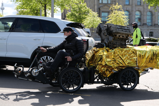 Ein Mann im Rollstuhl mit einem großen Motor an der Rückseite, umgeben von Fahrzeugen auf einer Straße mit Bäumen, Gebäuden und einem klaren blauen Himmel im Hintergrund; er trägt eine schwarze Jacke, eine Kappe und hält ein Objekt in der Hand.