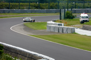 Ein Rennwagen fährt eine Rennstrecke entlang mit einem Polizeiwagen im Hintergrund, umgeben von Gras, Pflanzen, Bäumen und Absperrungen, mit einem Fahrzeug auf der rechten Seite und Text unten.