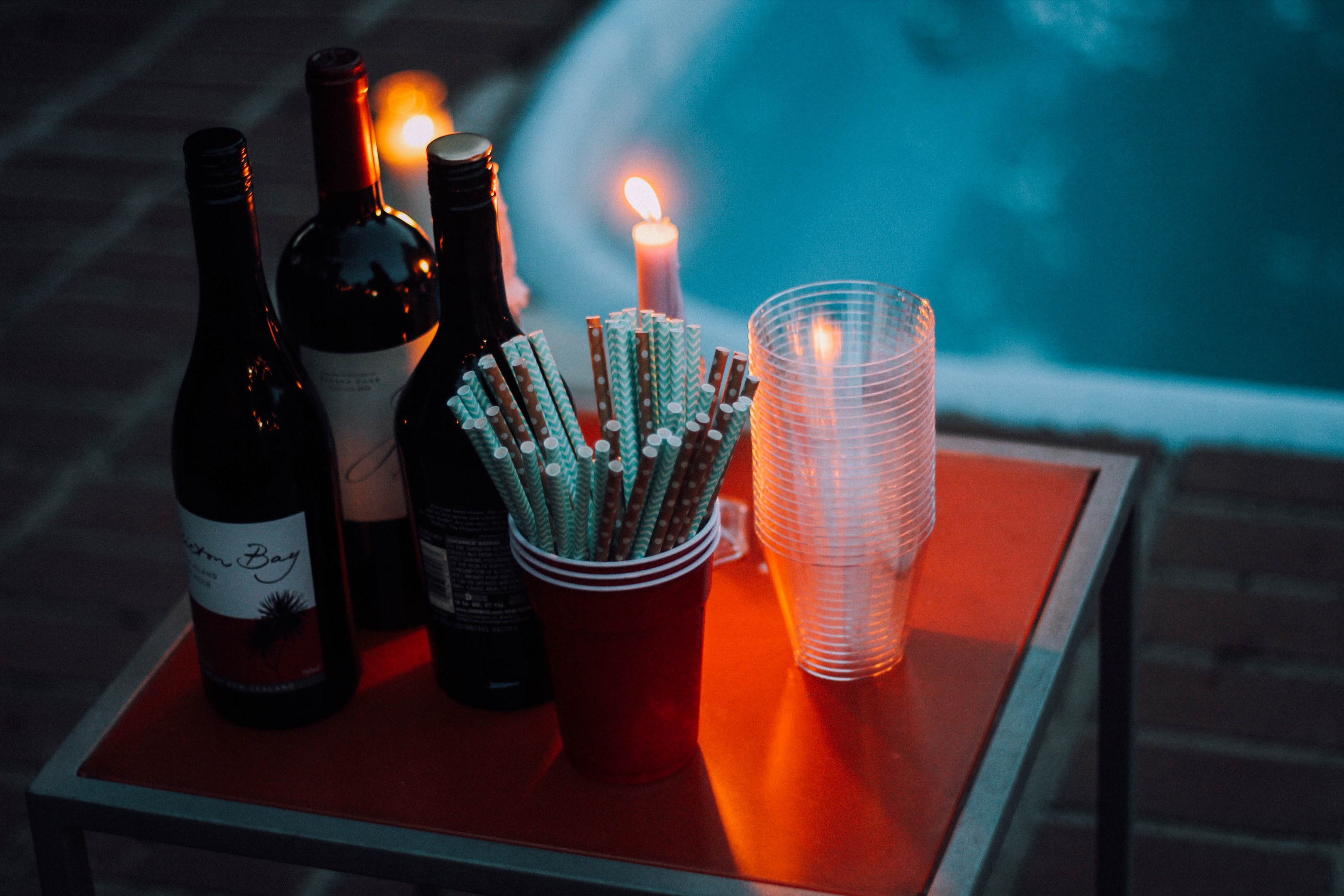 Ein Tisch am Swimmingpool mit zwei Weinflaschen, einer Kerze, Gläsern und Strohhalmen vermittelt Entspannung oder einen Abendausgang.