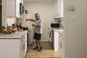 Ein Mann steht in einer Küche mit weißen Schränken, hält ein Glas in der Hand und verschiedene Gegenstände wie Obst, Gemüse, Schüsseln und Löffel sind auf einer Arbeitsplatte links von ihm.