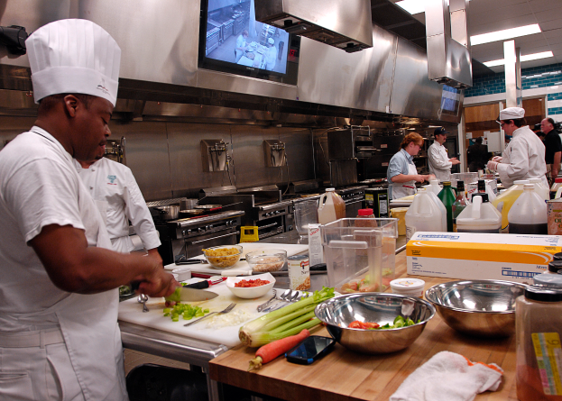 Eine Gruppe von Köchen in einer Küche, die auf einem Schneidbrett Essen zubereiten, mit einem Koch im Vordergrund in weißer Kleidung und Mütze, der ein Messer hält und von Utensilien, Schüsseln, Gemüse, einem Handy, Flaschen und anderen Gegenständen umgeben ist, mit einem Fernseher und Deckenleuchten im Hintergrund.