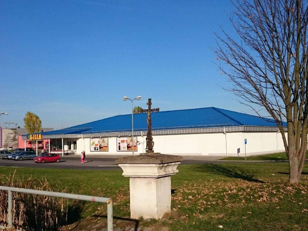 Ein große Gebäude mit einem blauen Dach, das als St. Paul's Catholic Church identifiziert ist, umgeben von einem Metallzaun, Gras, trockenen Blättern, Bäumen, Fahrzeugen, Menschen auf einem Fußweg, Masten, Schildern, einer Fahne mit Text, anderen Gebäuden mit Fenstern und einem bewölktem Himmel.