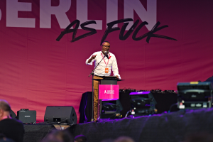 Ein Mann in einem wei├čen Hemd und braunen Hosen steht auf einer Bühne und h├Ąlt ein Mikrofon in der Hand, vor einem pinken Banner mit der Aufschrift "Berlin as Fuck" und sichtbarer Bühnenausrüstung.