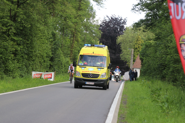 Ein Rettungswagen fährt auf einer Straße mit Radfahrern daneben, Gras und Bäume auf beiden Seiten, Häuser und Masten im Hintergrund unter einem klaren blauen Himmel.