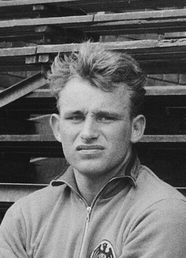 Schwarzes und weißes Foto eines Mannes in einer Jacke, der vor gestapelten Holzplanken steht, der als legendärer deutscher Fußballspieler gilt.