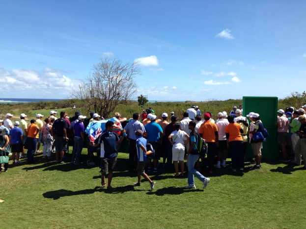Gruppe von Menschen auf einem grünen Feld bei der Beobachtung eines Golfturniers, mit Bäumen, Pflanzen, Wasser und wolkenverhangenem Himmel im Hintergrund.
