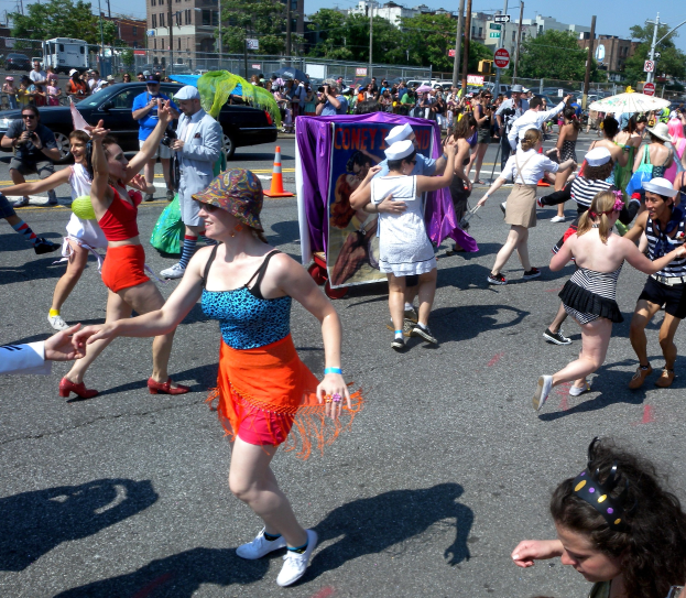 Eine Gruppe von Menschen, die eine Straße entlangmarschieren, einige mit Mötzen und Schirmen, andere tanzen, mit einem Banner, Verkehrskegeln, Fahrzeugen, Laternenmästen, Schildern, Bäumen, Gebäuden und einem klaren blauen Himmel im Hintergrund.