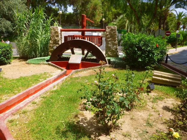 Minigolfanlage in einem Park mit Grünfläche, einer Straße mit Fußgängern und Hinweisschildern sowie einem blauen Himmel im Hintergrund.