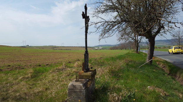 Ein gelbes Auto fährt an einem Baum und einem Straßenkreuz vorbei, mit Hügeln, einem bewölkten Himmel, Gras und Blumen im Hintergrund.