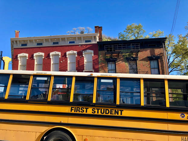 Ein gelber Schulbus der Marke "First Student" vor einem roten Backsteingebäude geparkt, mit ein paar Menschen drinnen und einem klaren blauen Himmel im Hintergrund.