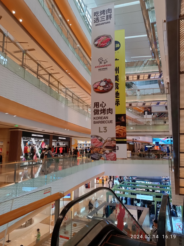 Ein Innenblick in ein Einkaufszentrum mit einer Rolltreppe in der Mitte, Menschen, die herumlaufen, einige mit Handtaschen, und Schilder mit Text an der Decke, mit Geschäften im Hintergrund.