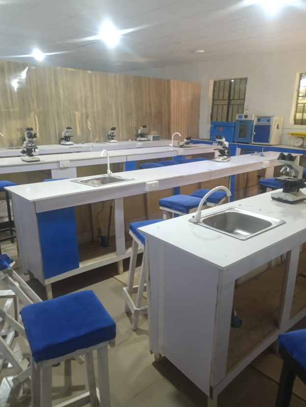 Ein Labor mit blauen Hockern und weißen Arbeitsplatten, einem Waschbecken mit Wasserhähnen, verschiedenen Maschinen, einer Wand mit Fenstern und Deckenleuchten, Teil der Universität für Wissenschaft und Technologie in Ahmedabad.