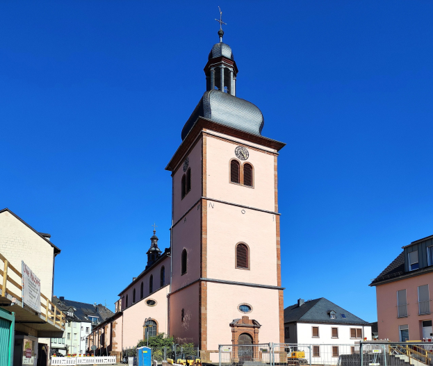Große rosafarbene Kirche mit einem zentralen Uhrenturm, umgeben von einem Metallzaun, in Baden-Württemberg, Deutschland.