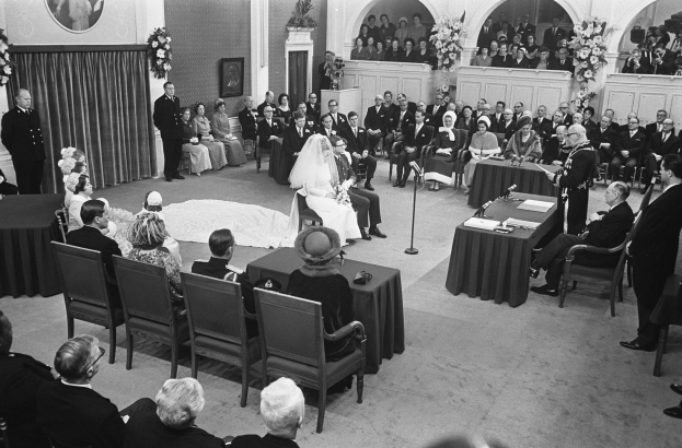 Schwarzes und weißes Foto einer Hochzeitszeremonie in einem großen Raum mit sitzenden und stehenden Gästen, Tischen mit Mikrofonen und Papieren, einer Wand mit einem Fotorahmen, Blumengestecken, einem Vorhang und einem Podium mit einem Mikrofonständer.