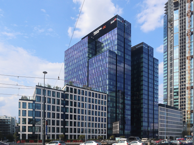 Zentrale der Deutschen Bank in Frankfurt, Deutschland, ein großes Gebäude mit vielen Fenstern, umgeben von städtischen Elementen einschließlich Straßenlaternen, Fahrzeugen und Bäumen unter einem bewölkten Himmel.