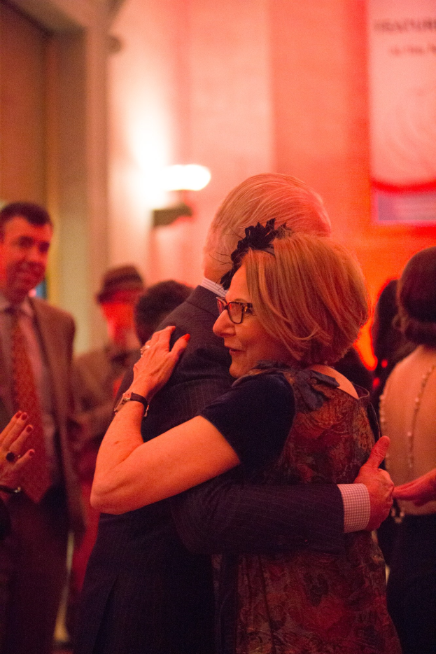 Ein Mann und eine Frau umarmen sich herzlich auf einer Party, umgeben von anderen Menschen und einer Wand mit einer Leuchte im Hintergrund.