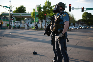 Ein Polizist in voller Montur, einschließlich einer Gasmaske, steht auf der Seite einer Straße und hält eine Waffe, mit Fahrzeugen, Verkehrszeichen, Schildern, Bäumen, Gebäuden und einer Tankstelle im Hintergrund.