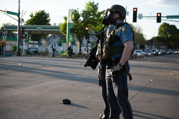 Ein Polizist in voller Montur, einschließlich einer Gasmaske, steht auf der Seite einer Straße und hält eine Waffe, mit Fahrzeugen, Verkehrszeichen, Schildern, Bäumen, Gebäuden und einer Tankstelle im Hintergrund.