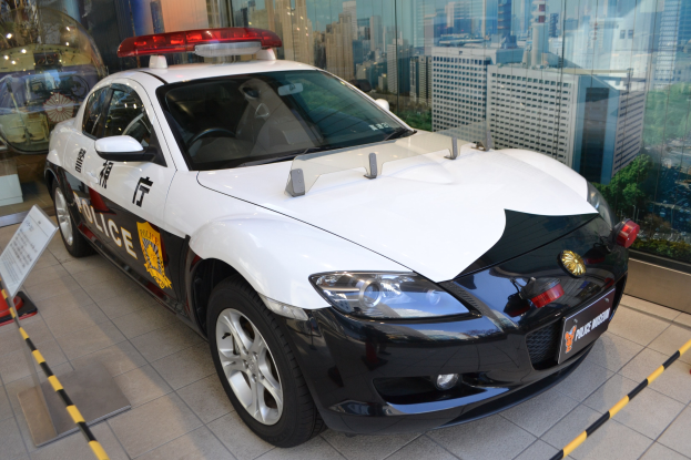 Polizeiauto vor einer Glaswand geparkt, mit sichtbarem Rad und einem Schild daneben, Gebäude, Bäume und Himmel durch die Glaswand sichtbar