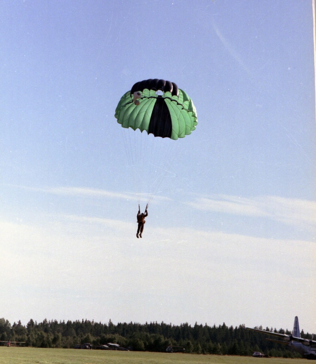 Eine Person beim Paragleiten in der Luft mit einem Fallschirm, mit einem Flugzeug rechts im Bild, vor einem Hintergrund aus Gras, Bäumen und wolkenbedecktem Himmel.