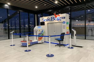 Sicherheitsbag-Check-in-Schalter am Flughafen mit einem Stuhl, Absperrpollern, Maschinen, einem Bildschirm, einer bedruckten Tafel, Glaswänden, Deckenleuchten und sichtbarem Boden.