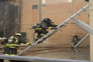 Feuerwehrleute in Helmen und Ausrüstung, die vor einem Backsteingebäude eine Leiter hochklettern, mit Rohren auf dem Boden und einer Metallstange am unteren Ende, mit einem weiteren Gebäude mit Fenstern und einem Netz im Hintergrund.
