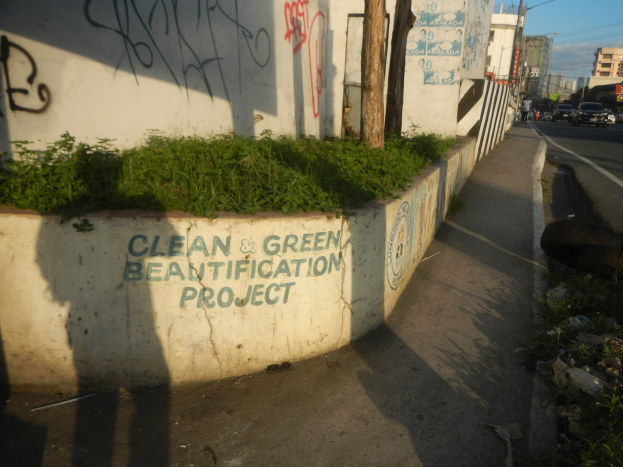 Eine Straße mit Fahrzeugen, Gebäuden, Bäumen, Pfählen, Drähten und einer Wand mit Graffiti "Saubere und grüne Verschönerung" umgeben von Pflanzen, Gras und einem bewölkten Himmel.