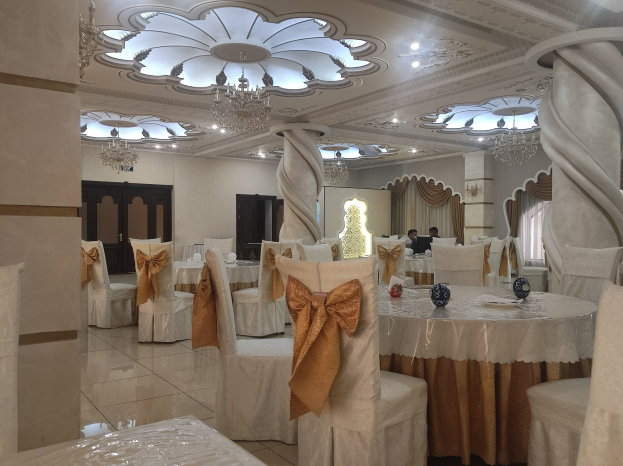 Ein großer Festsaal mit Tischen und Stühlen, die mit Tüchern bedeckt und mit Dekorationsgegenständen geschmückt sind, mit Türen und Vorhängen an den Wänden und beleuchteten Kronleuchtern an der Decke.
